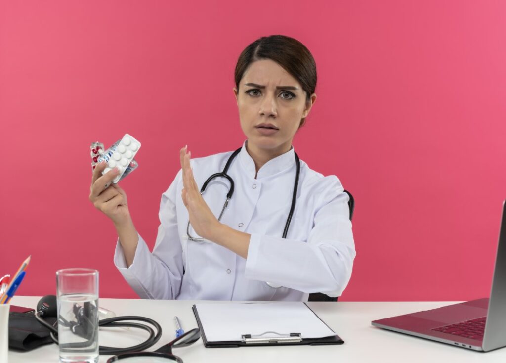 frowning-young-female-doctor-wearing-medical-robe-stethoscope-sitting-desk-with-medical-tools-laptop-holding-medical-drugs-gesturing-no-isolated-pink-wall